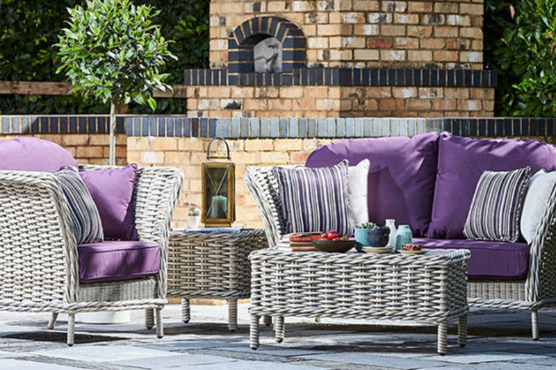 Outdoor rattan patio furniture set with purple cushions in front of a brick wall.